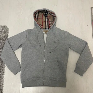Grå hoodie från Burberry - Säljer en stilren grå hoodie från Burberry med klassiskt rutigt foder i huvan. Tröjan har dragkedja framtill och ribbade muddar vid ärmslut och nederkant. Perfekt för en avslappnad och trendig look.