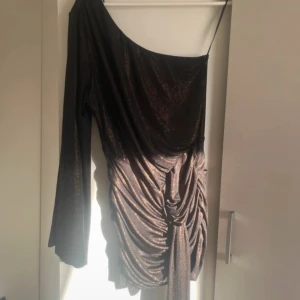 Glittrig brun one-shoulder klänning - Snygg glittrig brun/guld klänning med one-shoulder design och långa ärmar. Klänningen har en draperad effekt med knytning i midjan som ger en elegant look. Perfekt för en kväll ute eller en speciell tillställning.