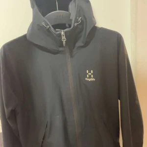 Svart jacka från Haglöfs - Säljer en stilren svart jacka från Haglöfs med dragkedja och huva. Perfekt för utomhusaktiviteter med sin funktionella design. Jackan har långa ärmar och en diskret logga på bröstet.