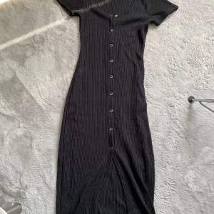 Svart ribbad klänning från H&M Divided - Snygg svart ribbad klänning från H&M Divided i storlek XS. Klänningen har korta ärmar och knappar längs hela framsidan. Perfekt för en stilren look.