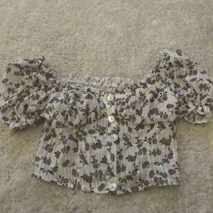 Blommig blus - Charmig blommig blus med volangdetaljer och knappar framtill. Blusen har ett romantiskt mönster i lila och grönt på en vit bakgrund. Perfekt för en somrig look!