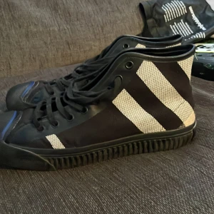 Svarta sneakers med vita ränder från Burberry - Snygga svarta sneakers från Burberry med vita ränder och en blå detalj på hälen. Skorna har snörning och en robust sula med Burberry-logga. Perfekta för en stilren look. Använda ett få gånger nypris 5500kr.