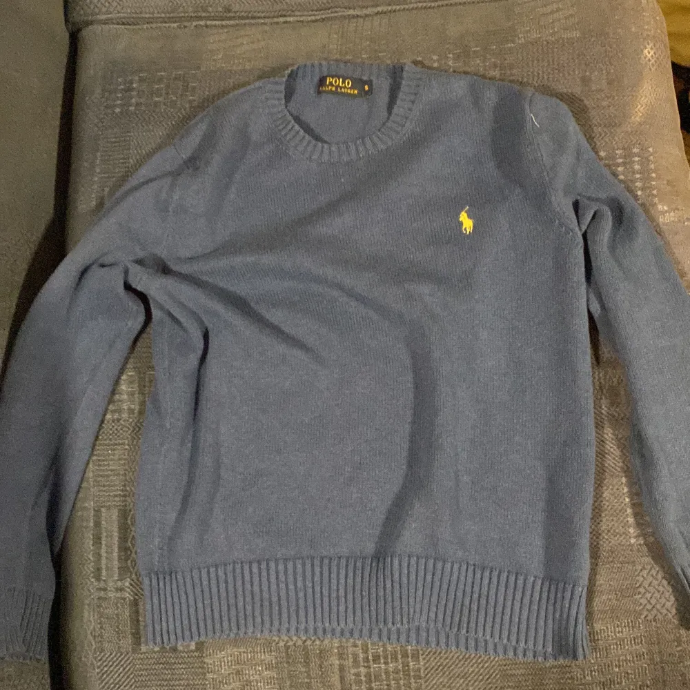 Säljer en stilren blå tröja från Polo Ralph Lauren med det klassiska gula logotypbroderiet på bröstet. Tröjan har långa ärmar och ribbade muddar vid ärmslut och nederkant. Perfekt för en avslappnad och snygg look.. Neuletakit & Villapaidat.