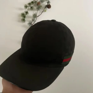 Svart gucci keps  - Snygg svart keps från Gucci med diskret mönster och klassiska ränder i grönt och rött på sidan. Perfekt för att ge din outfit en lyxig touch. Justerbar passform för optimal komfort.