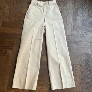Beige vida byxor - Snygga beige byxor med hög midja och vida ben. Perfekta för en stilren look. Byxorna har en klassisk design med knappar och dragkedja framtill. Har en liten defekt vid bakfickan. Liten i storleken 32/34 skulle jag säga. 