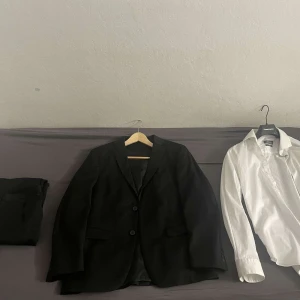 Svart kostym från Dressmann - Säljer en stilren svart kostym från Dressmann i storlek 48. Kostymen har en kavaj med två knappar och matchande byxor. Byxorna är storlek 33/30. Perfekt för formella tillfällen. Skjortan är vit och långärmad, vilket kompletterar kostymen snyggt. Skjortan är storlek M. För fler bilder är det bara att höra av sig!
