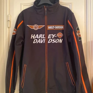 Svart Harley-Davidson jacka med broderier - Snygg svart Harley-Davidson jacka med orange detaljer och flera broderade logotyper. Jackan har dragkedja framtill och långa ärmar. Perfekt för motorcykelentusiaster!