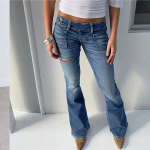 HELT NYA GINA JEANS lappar kvar! - Helt nya med prislapp snygga blå bootcut jeans med slitningar på låret. De har en låg midja och dubbla knappar i midjan för en cool look. Perfekta för en avslappnad stil.