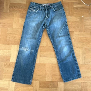 Diesel Jeans - Säljer ett par klassiska blå jeans från Diesel i straight passform. Jeansen har en sliten detalj på ena benet och fem fickor med knapp och dragkedja i gylfen. 