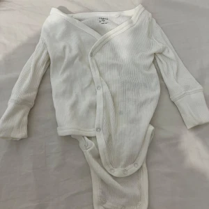 Vit ribbad babybody från Lindex - Säljer en söt vit ribbad babybody från Lindex i storlek newborn. Bodyn har långa ärmar och praktiska knappar för enkel på- och avklädning. Perfekt för de allra minsta! 🌟