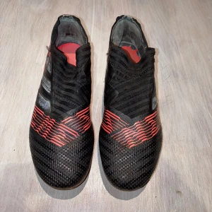 Svarta och röda fotbollsskor från Adidas - Snygga fotbollsskor från Adidas i svart med röda detaljer och guldiga sulor. Skorna har en strömlinjeformad design utan snörning för en tight passform. Perfekta för spel på gräsplaner.