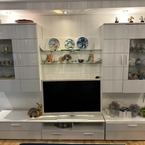 Vit väggmonterad TV-bänk med vitrinskåp - Elegant vit väggmonterad TV-bänk med integrerade vitrinskåp och hyllor. Perfekt för att visa upp prydnadssaker och förvara elektronik. Modern design med glasdörrar och inbyggd belysning för en stilren look.