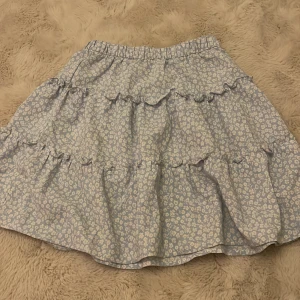 Blommig kjol från H&M - Söt blommig kjol från H&M i ljusblått med vita blommor. Kjolen har volanger och en elastisk midja för en bekväm passform. Perfekt för en somrig look!