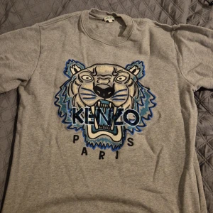 Grå sweatshirt från Kenzo - Säljer en grå sweatshirt från Kenzo med ett stort tigertryck och texten 'Kenzo Paris' på framsidan. Tröjan har rund hals och långa ärmar. Perfekt för en avslappnad stil. Kan sänka priset.