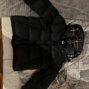 Svart pufferjacka från Burberry - Säljer en snygg svart pufferjacka från Burberry. Jackan har avtagbara ärmar och en klassisk Burberry-rutig insida i huvan. Den är utrustad med dragkedja och har en stilren design med märkets logga på ärmen. Jackan är i storlek S men den sitter som storlek M. Med  avtagbara ärmar kan du enkelt anpassa den efter vädret och din stil.
