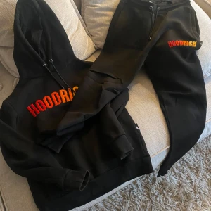 Svart hoodie set från Hoodrich - Säljer en svart hoodie från Hoodrich med färgglad logga i rött och gult på bröstet. Hoodien har en klassisk design med dragsko i huvan och ribbade muddar vid ärmslut och nederkant. Perfekt för en avslappnad stil. Byxor ingår