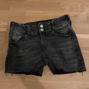 Svarta jeansshorts med fickor i baksidan - Säljer ett par svarta jeansshorts med fransiga kanter och dubbla knappar fram. Shortsen har fickor både fram och bak, vilket ger en cool och avslappnad look. Perfekta för en somrig dag eller en avslappnad kväll.