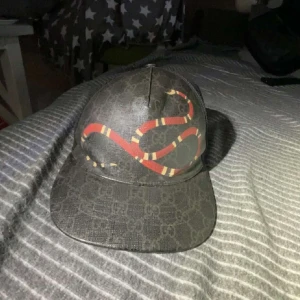 Gucci keps - Snygg svart keps med ett iögonfallande ormmönster i rött och gult. Kepsen har en justerbar baksida och ett diskret mönster i tyget. Perfekt för att ge din outfit en cool touch.
