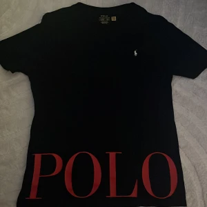 Svart t-shirt från Ralph Lauren - Snygg svart t-shirt från Ralph Lauren med stor röd 'POLO' text på framsidan och det klassiska broderade logotypen på bröstet. Perfekt för en stilren look, den sitter som M  (Aldrig använts helt ny). Skriv för snabb affär om priset eller funderingar vill bara få den sålt kommer ej till användning.