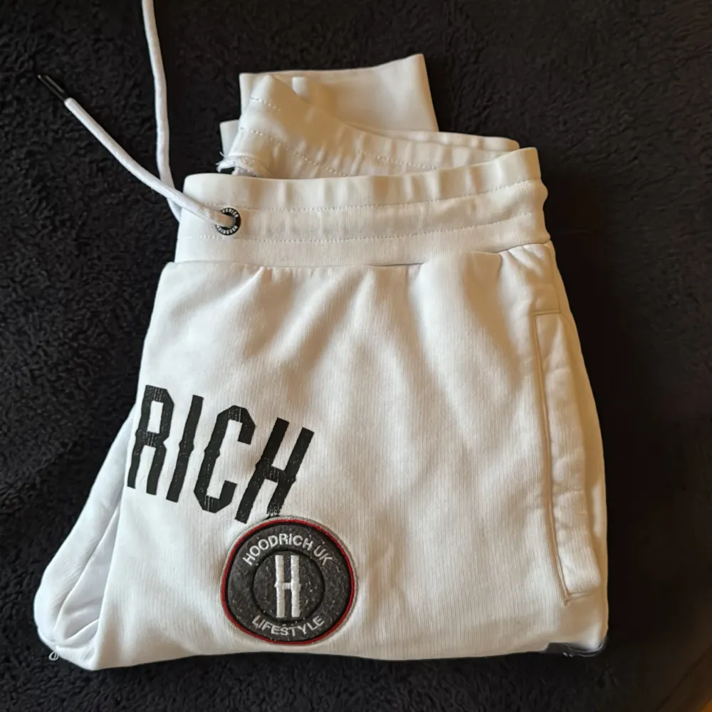 Säljer ett par vita mjukisbyxor från Hoodrich med snygga broderade detaljer och logotyper. Byxorna har snörning i midjan och är perfekta för en avslappnad stil. De är tillverkade i ett mjukt material som ger en bekväm passform.. Farkut & Housut.