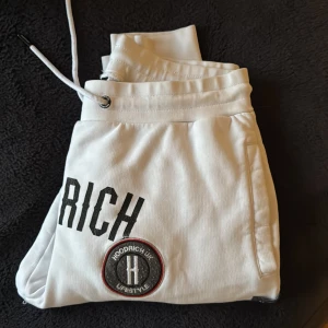 Vita mjukisbyxor från Hoodrich - Säljer ett par vita mjukisbyxor från Hoodrich med snygga broderade detaljer och logotyper. Byxorna har snörning i midjan och är perfekta för en avslappnad stil. De är tillverkade i ett mjukt material som ger en bekväm passform.