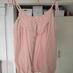 Rosa topp från H&M - Säljer en söt rosa linnetopp från H&M med knappar framtill och volangdetaljer vid halsen. Toppen har justerbara axelband som knyts och en luftig passform. Perfekt för varma dagar!