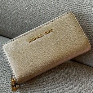 Guld plånbok från Michael Kors - Snygg guldplånbok från Michael Kors med dragkedja runtom. Plånboken har flera fack för kort och sedlar, perfekt för att hålla ordning på dina tillhörigheter. Den eleganta designen gör den till ett stilrent val för alla tillfällen.