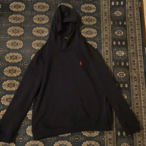 Svart hoodie från Polo Ralph Lauren - Säljer en stilren svart hoodie från Polo Ralph Lauren med det klassiska röda logotypen på bröstet. Tröjan har en bekväm passform och en praktisk huva. Perfekt för en avslappnad look.