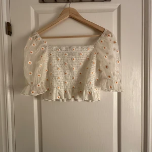 Vit blus med blommönster från Bik Bok - Charmig vit blus från Bik Bok med broderade blommor i gult och orange. Blusen har puffiga, transparenta ärmar och en smockad kropp med volangdetalj vid nederkanten. Perfekt för en somrig look! 🌼