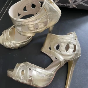 Högklackade skor med utskärningar 37 nya  - Snygga guldiga klackskor med unika utskärningar och öppen tå. Perfekta för att ge en glamorös touch till din outfit. Skorna har en elegant design med hög klack och dragkedja baktill för enkel på- och avtagning. Oanvända.  