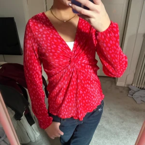 Röd mönstrad blus från JDY - Säljer en snygg röd blus från JDY med ett diskret mönster och v-ringning. Blusen har långa ärmar och en knutdetalj framtill som ger en fin siluett. Perfekt för att liva upp garderoben!