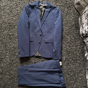 Marinblå/mörkblå kostym från Jack and Jones - En snygg kostym från Jack and Jones i perfekt skick bara använd några fåtal gånger inga defekter eller andra problem. Orginal pris 1200 kr storlek S barn rekommenderad storlek 13 år gammal/160 cm lång men funkar för lite längre också. Du får med en hel kostym alltså kostymbyxor och kavaj som du ser på bilderna. Det är bara och skriva vid funderingar.