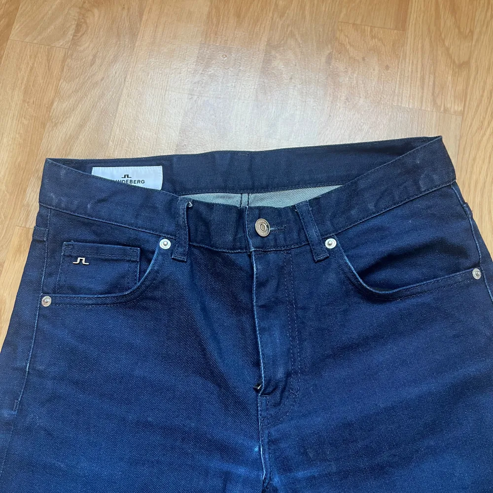 Snygga mörkblå jeans från J.Lindeberg med klassisk femficksdesign och knappgylf. Perfekta för en stilren look. Märkeslogga på bakfickan ger en extra touch.. Farkut & Housut.