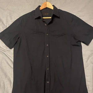 Svart kortärmad skjorta från Massimo Dutti - Säljer en stilren svart kortärmad skjorta från Massimo Dutti. Skjortan har en klassisk krage och knappar framtill. Perfekt för en elegant och avslappnad look.