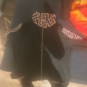 Svart jacka med mönstrade detaljer - Snygg svart jacka med unika mönstrade detaljer i beige runt kragen och ärmarna. Jackan har en dragkedja framtill och en stilren design som passar perfekt för både vardag och speciella tillfällen.