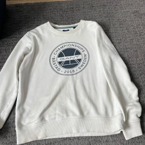 Vit sweatshirt från GANT - Säljer en stilren vit sweatshirt från GANT med ett tryck på framsidan som refererar till Båstad Championships 2018. Tröjan har långa ärmar och ribbade muddar vid ärmslut och nederkant. Perfekt för en avslappnad stil! Snygg överallt 