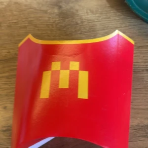 Röd pommes frites-förpackning med Minecraft-tema - En röd pommes frites-förpackning från McDonald's med ett Minecraft-tema. Förpackningen har en gul dekorativ kant och ett gult mönster på framsidan. Baksidan visar Minecraft-logotypen och en illustration. Perfekt för samlare eller Minecraft-fans!