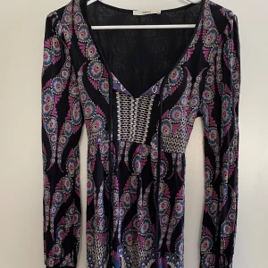 Mönstrad klänning från Oasis - Säljer en färgglad klänning från Oasis med ett unikt mönster i lila, rosa och blå nyanser på en svart bakgrund. Klänningen har långa ärmar och en v-ringad design. Perfekt för att sticka ut med stil!