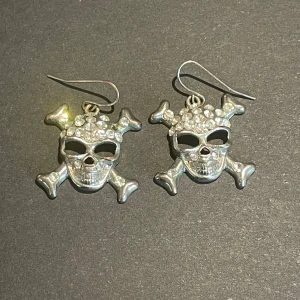 Silverfärgade dödskalleörhängen - Coola silverfärgade örhängen i form av dödskallar med korslagda benknotor. Dödskallarna är dekorerade med glittriga stenar för en extra edgy look. Perfekta dig som gillar alternativa smycken och kläder