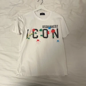 Vit t-shirt från Dsquared2 - Säljer en vit t-shirt från Dsquared2 med färgglada stänk och texten 'ICON' på framsidan. T-shirten har en normal passform och är kortärmad, perfekt för en avslappnad stil. Pris sänks vid snabbaffär.