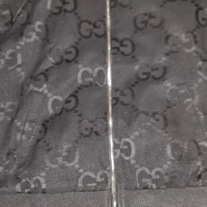 Svart mönstrad jacka med dragkedja - Snygg svart jacka med ett distinkt mönster och dragkedja framtill. Jackan har ribbade detaljer och en rand i grönt och rött längs sidan. Perfekt för en stilren look.