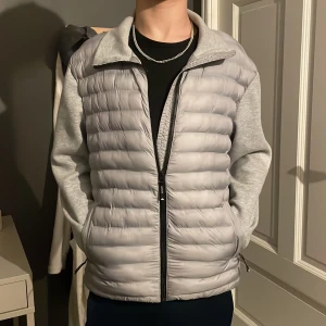 Grå dunväst med ärmar från Superdry - Snygg grå dunväst med ärmar från Superdry. Västen har en dragkedja framtill och är quiltad för extra värme. Perfekt för kyliga dagar med sin stilrena design och bekväma passform. Knappat använd och den passar 164-170