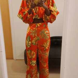 Färgglada blommiga byxor - Säljer ett par färgglada byxor med tropiskt blommönster i orange, grönt och gult. Byxorna har en hög midja och vida ben, vilket ger en avslappnad och trendig look. Perfekta för att liva upp garderoben!