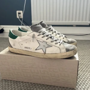 Golden Goose  - Snygga Golden Goose Super-Star sneakers i vitt med grå detaljer och en stjärna på sidan. Skorna har gröna hälpartier och en klassisk snörning. Perfekta för en avslappnad stil med en touch av lyx. Skorna är i fint skick och är använt sparsamt. Pris är ej hugget i sten, storlek 44