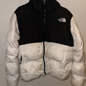 Svart och vit dunjacka från The North Face - Säljer en snygg svart och vit dunjacka från The North Face. Jackan har en dragkedja framtill och är långärmad. Perfekt för kyliga dagar med sin pufferdesign som håller dig varm och bekväm.