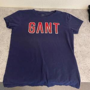 Mörkblå t-shirt från GANT - Säljer en snygg mörkblå t-shirt från GANT med röd och vit logga på framsidan. T-shirten är kortärmad och gjord i mjuk materiall. Hör av dig vid intresse!
