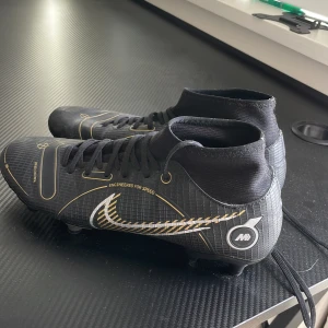 Svarta fotbollsskor med gulddetaljer - Snygga svarta fotbollsskor med gulddetaljer och snörning. Designade för hastighet och kontroll på planen. Perfekta för den som vill ha stil och funktion i ett.