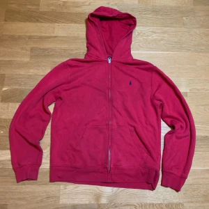 Röd hoodie från Ralph Lauren - Säljer en snygg röd hoodie från Ralph Lauren med dragkedja och klassisk logga på bröstet. Perfekt för en avslappnad stil. Den har en praktisk huva och fickor framtill. Den har två små hål som man knappt märker. Xl i barnstorlek