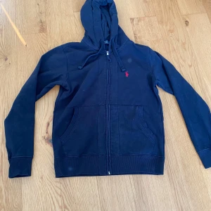 Svart hoodie från Polo Ralph Lauren - Den ser lite fläckig ut på bilden men det är bara min kamera. Säljer en marinblå hoodie från Polo Ralph Lauren i 100% bomull. Den har en dragkedja framtill och en liten röd logga på bröstet. Perfekt för en avslappnad stil. Inga defekter
