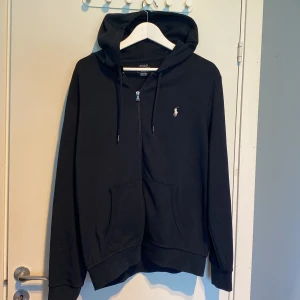 Blå hoodie från Polo Ralph Lauren - Säljer en stilren svart hoodie från Polo Ralph Lauren med dragkedja och broderad logotyp på bröstet. Tröjan har långa ärmar och en praktisk huva med snörning. Perfekt för en avslappnad stil.